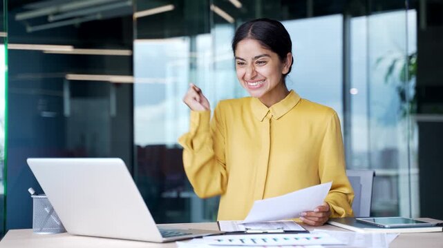 Happy young businesswoman sitting at desk in modern office looking at computer and financial document. Smiling female financier celebrating success, feeling satisfied with positive results and profit 