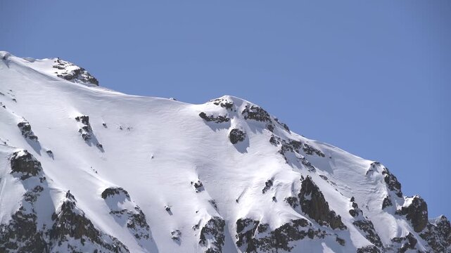 Panoramic snowy mountain range shows jagged rocky peaks, ridgeline contours, clear sky. Wide cinema vista reveals ice covered highland summits, crags, crest forms, azure backdrop.