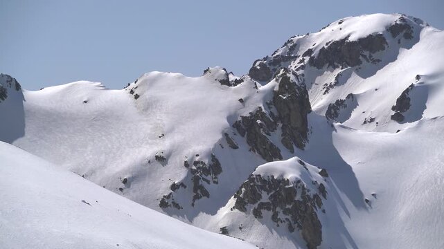 Panoramic snowy mountain range shows jagged rocky peaks, ridgeline contours, clear sky. Wide cinema vista reveals ice covered highland summits, crags, crest forms, azure backdrop.