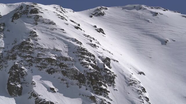 Panoramic snowy mountain range shows jagged rocky peaks, ridgeline contours, clear sky. Wide cinema vista reveals ice covered highland summits, crags, crest forms, azure backdrop.
