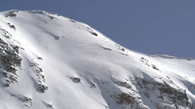 Panoramic snowy mountain range shows jagged rocky peaks, ridgeline contours, clear sky. Wide cinema vista reveals ice covered highland summits, crags, crest forms, azure backdrop.