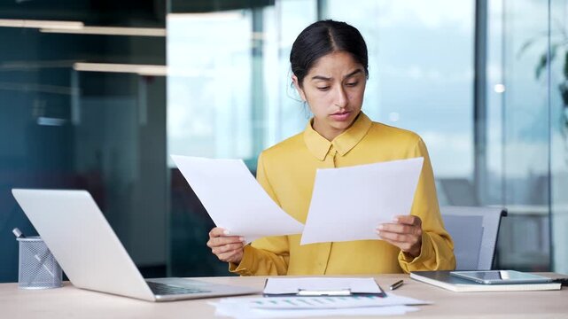 Confused young businesswoman sitting at desk in modern office holding documents in hands. Frustrated female employee having difficulty with paperwork, looking at papers in disbelief and uncertainty.