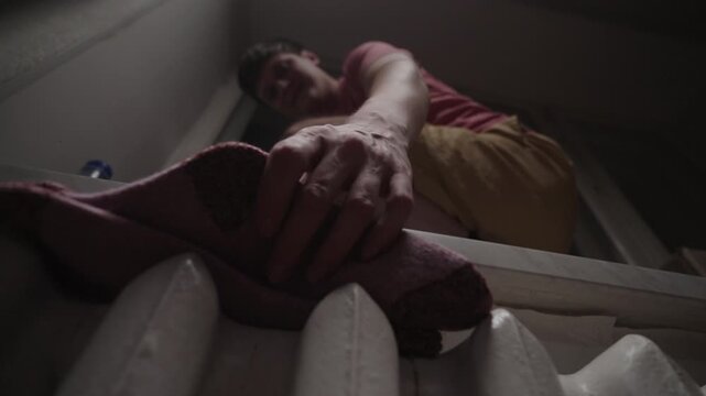 Young man sitting on windowsill placing wet insoles and socks on warm radiator to dry, giving positive thumbs-up gesture, symbolizing resourcefulness during energy crisis, close-up, slow motion.