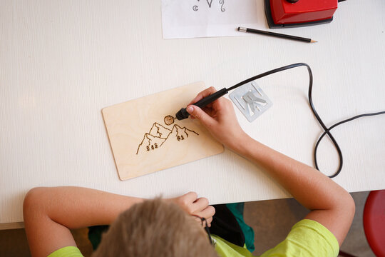 A child is using a pyrography pen to burn a mountain design onto a wooden board. Pencil, paper with sketches and a red pyrography tool are visible on the table - a creative craft session