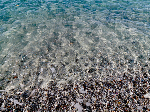 Clear turquoise ocean water gently flows over the pebbly bottom under bright sunlight, creating shimmering light patterns on the shallow shore, close-up