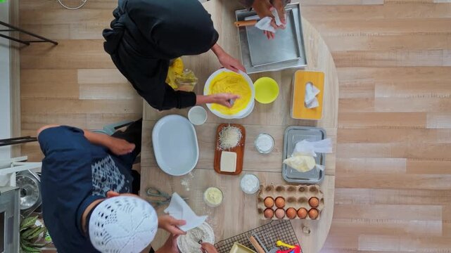 Top view of family preparing traditional Eid cookies mixing yellow cookie batter and organizing baking ingredients on wooden table 