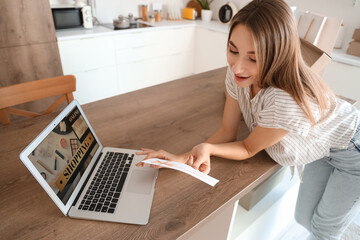 Young woman with gift card and laptop shopping online in kitchen