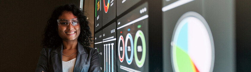 A confident Indian businesswoman stands in front of glowing digital screens with graphs and data...