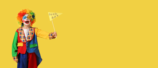Obraz premium Funny little boy in clown costume with paper flag on yellow background. April Fools' Day celebration