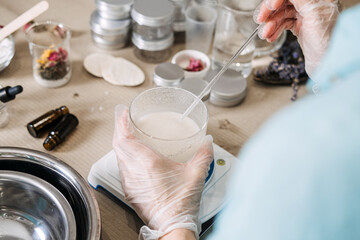 Hands in gloves use glass rod to stir white liquid in beaker on digital scale. Product transformation, raw to ready, artisanal skincare journey, behind the brand.