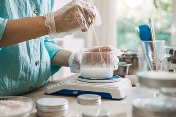 Person uses glass rod to stir white liquid in glass beaker on digital scale. Molecular skincare formulation, active ingredient dissolving, functional beauty craft, precision cosmetic measurement.