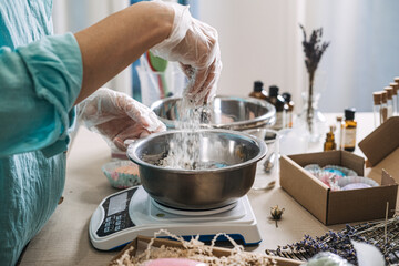 Person sprinkles white powder into metal bowl on digital scale for cosmetic preparation. Floral bath salt layers, apothecary bath mixology, botanical soak formulation, aesthetic dry ingredients mix.