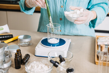 Person uses green spatula to transfer white powder from bowl into glass beaker on digital scale. Clean beauty science, eco-friendly lab, natural formulation expert, sustainable beauty craft.