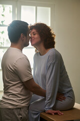 Fototapeta premium Vertical shot of man embracing woman sitting on table, both smiling with eyes closed, man gently touching woman's waist, intimate moment in bright room