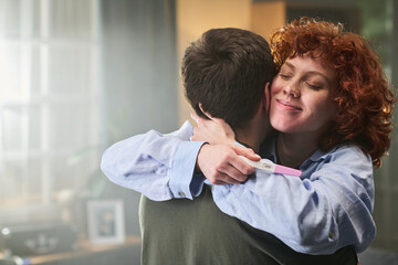 Fototapeta premium Caucasian young adult woman hugging Caucasian young adult man while holding positive pregnancy test, smiling with eyes closed, standing indoors in home environment