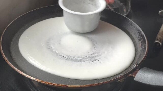 Side angle close-up of spreading dosa batter into a perfect round shape on hot tawa, traditional South Indian cooking in home kitchen.