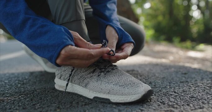 Ground level view shows a woman kneeling, tightening black and white laces on a light gray SPORT running shoe in a public park with golden evening light and bokeh.