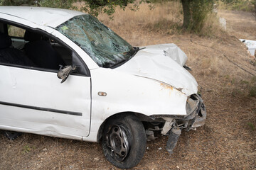 Crashed white car abandoned in dry roadside grass, showing shattered windshield, a missing front bumper, and a flat tire