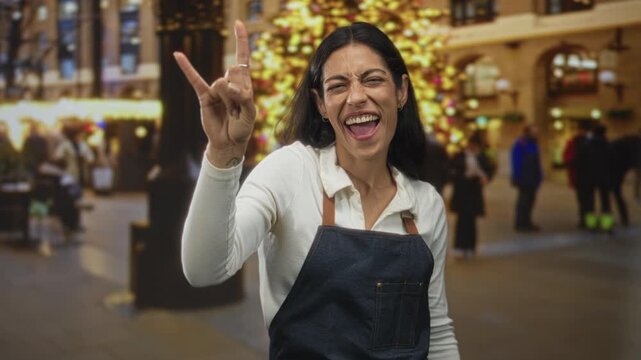 Woman in denim apron flashing rock horns gesture on street with holiday lights and blurred crowd; joy celebration.