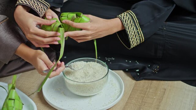 Close-up of two muslim women hands using spoon to fill ketupat shells with rice during Lebaran preparations
