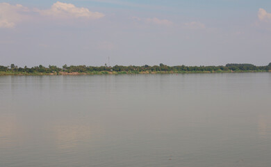 Naklejka premium Vast river flows calmly past a lush green tropical shoreline under a soft, hazy summer sky.