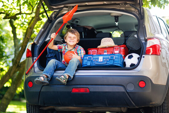 Adorable little kid boy sitting in car trunk just before leaving for summer vacation with his parents. Happy child with suitcases and toys going on journey, real life, real people