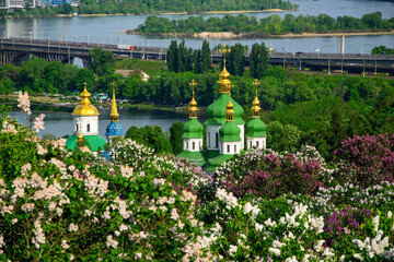 Vydubychi Monastery with green and golden domes framed by blooming lilacs, overlooking the Dnieper and a distant bridge on a bright spring day in Kyiv, Ukraine © haidamac