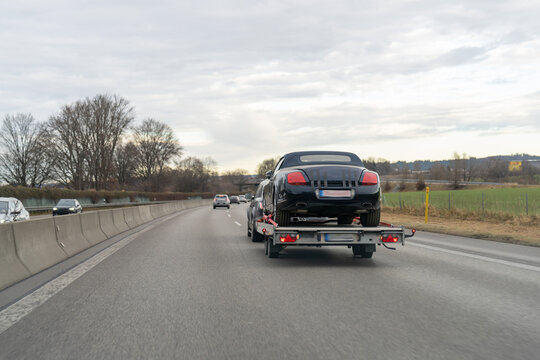 Black Bentley convertible car transported on a flatbed trailer along a European highway on a cloudy winter day