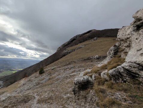 Paesaggio montano con grandi formazioni rocciose in primo piano, pendio erboso e vista sulla valle sotto un cielo nuvoloso