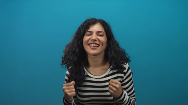 Woman in striped shirt clenching fists with head tilted back and exposed neck in a teal studio; excitement.