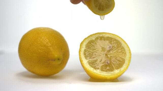 Slow motion horizontal video of a hand squeezing one half of a lemon over another sliced half resting on the surface, with a whole lemon placed beside