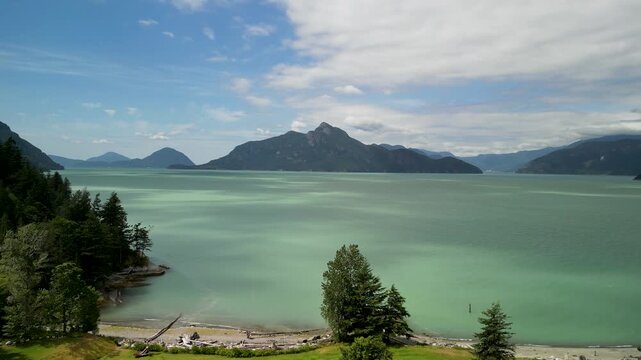 A drone captures turquoise glacial waters meeting lush green shoreline and distant mountains under a partly cloudy sky. Calm water shimmers while grassy fields, small trees and rugged peaks create a t