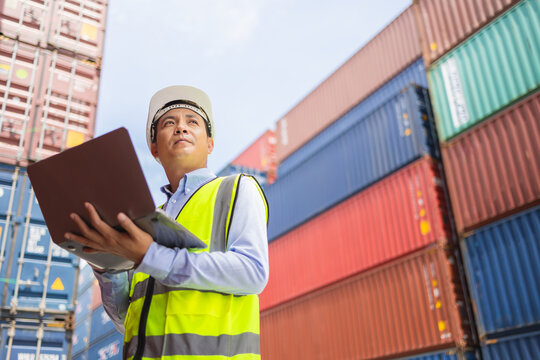 Engineer wearing safety vest and helmet using laptop to inspect cargo containers at shipping yard, managing logistics and cargo operations in industrial transportation concept.