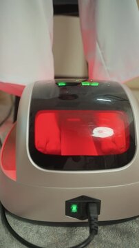 red lit foot device on carpet, glossy control panel with green power indicator, plugged cord and reflective window, sheer curtain backdrop, ambient clinical lighting, microcurrent therapy unit