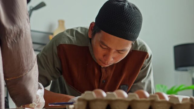 Focused Indonesian man wearing kufi hat making traditional Eid treats with family
