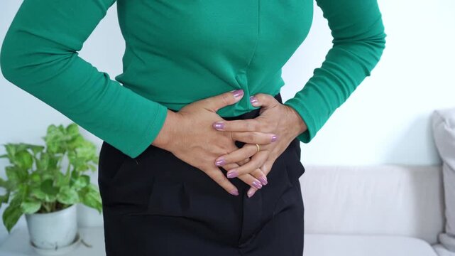 close-up of a woman clutching her stomach in pain, depicting abdominal cramps, indigestion, or menstrual discomfort while standing in a living room. Medical health concept