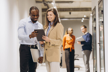 Obraz premium Smiling business partners using digital tablet and discussing work while standing in office hallway with colleagues working in background