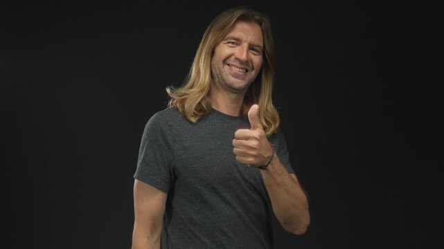 Smiling man with long blond hair gives thumbs up in studio with black background and friendly gaze; optimism.