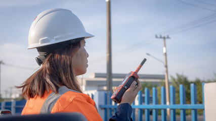 A person stands at a construction site wearing a helmet and an orange jacket. They hold a walkie-talkie and talk, looking towards the distance © Happy Photo