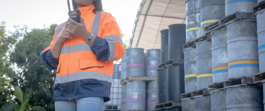 A person wearing a bright safety jacket and jeans stands near stacked barrels in a storage area. The worker is using a two-way radio to communicate while observing the surroundings