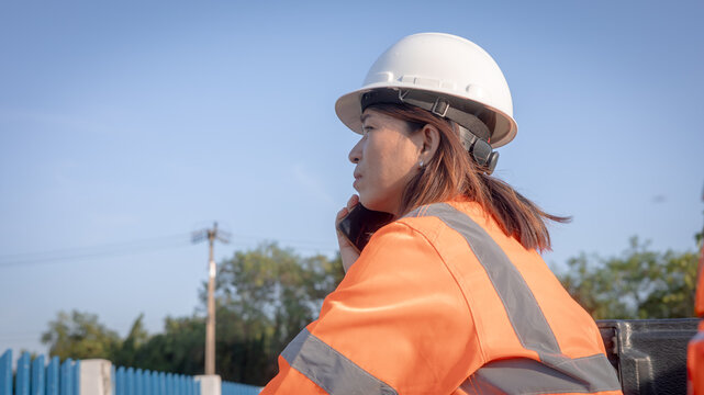 A construction worker stands in a work area wearing a safety helmet and orange jacket while talking on a mobile phone. It is a clear sunny day, and trees are visible