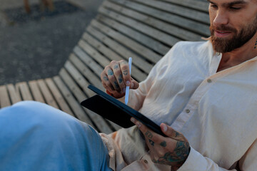 Casual man with tattoos using a tablet and stylus on a wooden bench outdoors in Lisbon