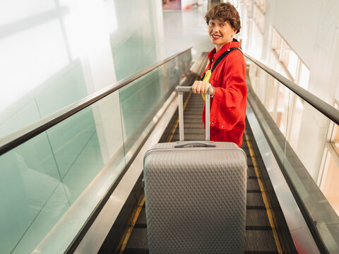 A young woman in red with a suitcase on an escalator at an airport or train station looks at the camera. Traveling with luggage.