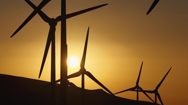 Aerial view of wind turbines silhouetted against a vibrant sunset, creating a striking contrast of dark shapes against the warm, glowing sky, Lanzarote, Spain.