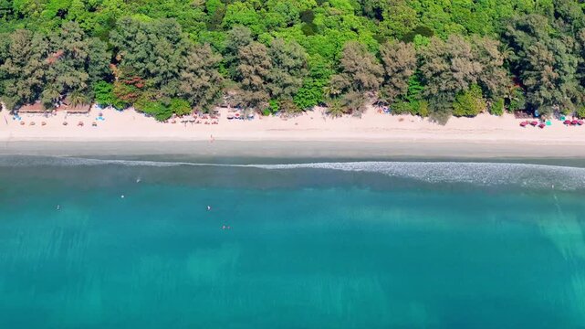 Top-down aerial view of Koh Phayam beach showing turquoise water, gentle waves, swimmers, surfers, and sandy shoreline bordered by trees.