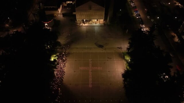 Drone view of worshippers carrying candles around a glowing cross shape. The evening procession fills the square with warm light and quiet devotion in the Milan suburb.