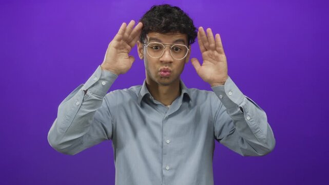 Young hispanic man with glasses covers eyes with open hands in purple studio setting; shyness privacy.