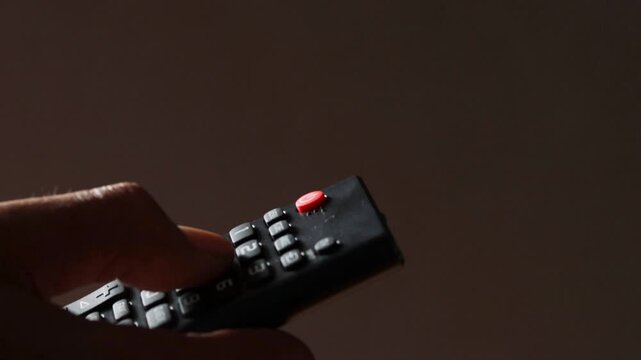 Unrecognizable male hands holding a TV remote control while switching TV channels in a dimly lit room.