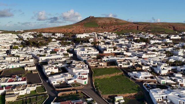 Aerial view of Teguise's white buildings contrasting with the reddish mountain backdrop under a blue sky, Teguise, Canary Islands, Spain.
