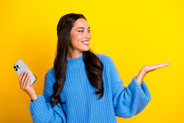 Young mixed race woman in blue sweater smiling holding smartphone against bright yellow background...